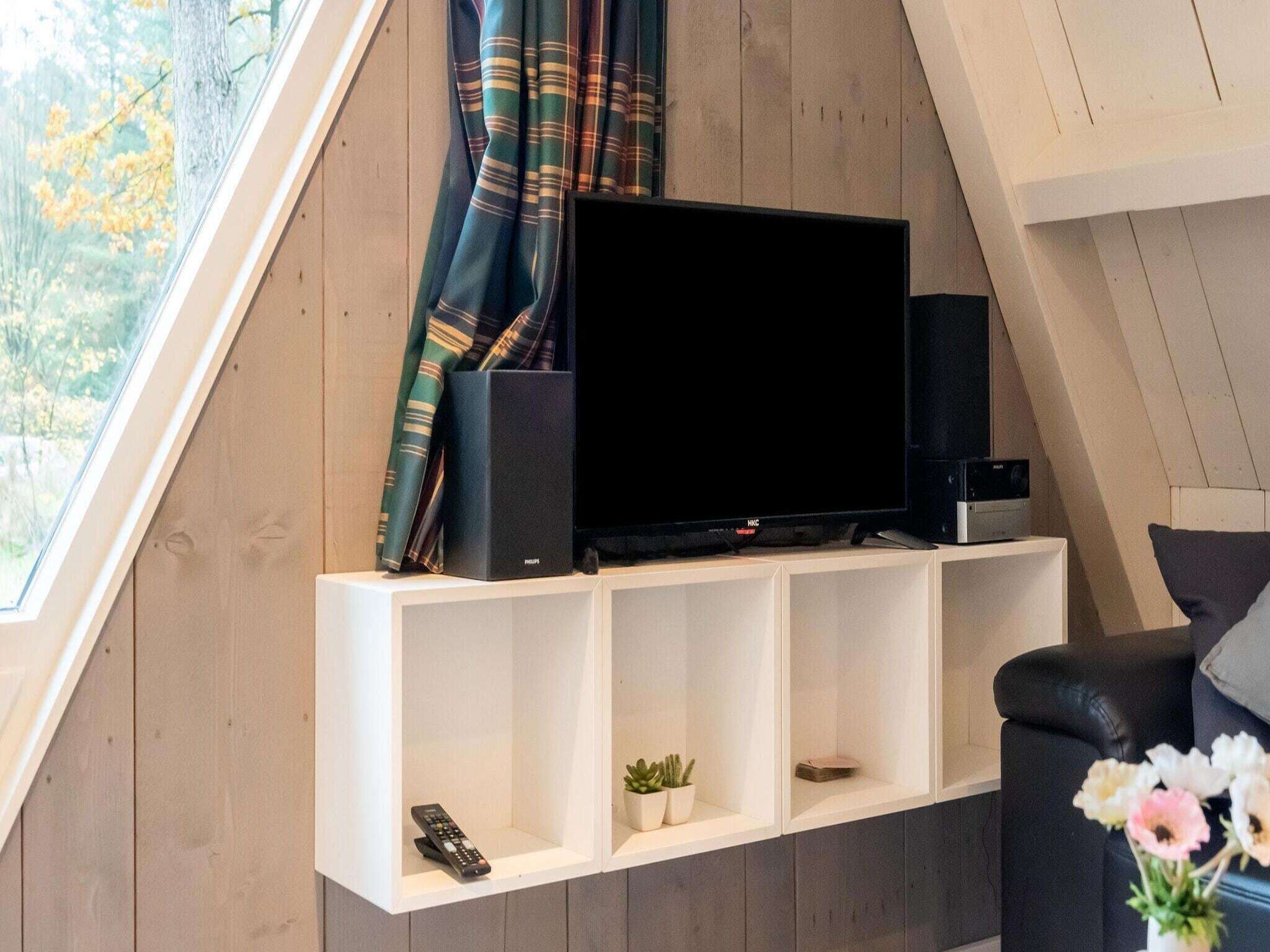 Ett modernt vardagsrum med en platt-TV på en vit hylla, en fjärrkontroll och en liten växt.