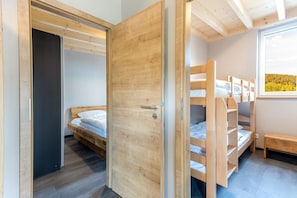 A room with a wooden door, a bunk bed, a small bench, and a window with a view of trees.
