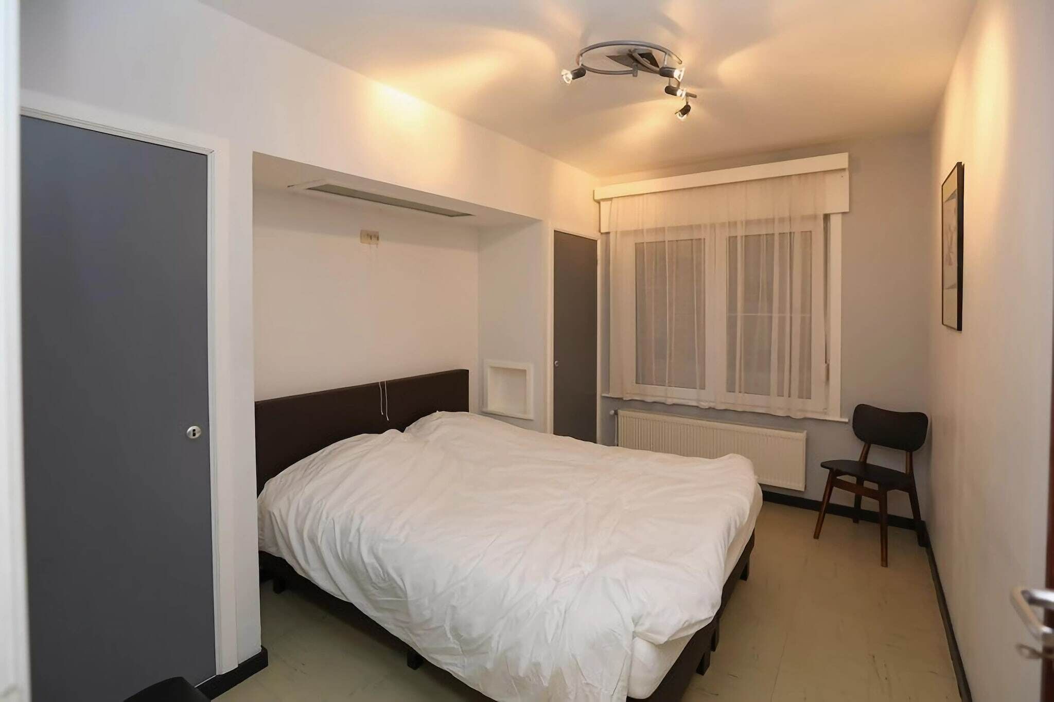 Ein Hotelzimmer mit Bett, Stuhl, einem Fenster mit Vorhängen, Heizkörper und einer Deckenleuchte.