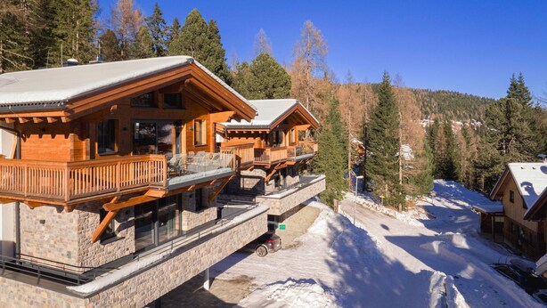 Un edificio in stile chalet con fondamenta in pietra e facciata in legno, dotato di balconi e ampie finestre, immerso in un paesaggio montano innevato.