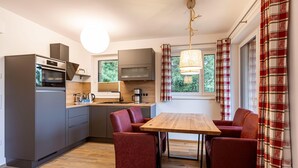 Una cucina compatta con elettrodomestici moderni, un'area pranzo con un tavolo di legno e sedie rosse, e ampie finestre con tende a quadri.