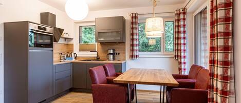 A compact kitchen with modern appliances, a dining area with a wooden table and red chairs, and large windows with plaid curtains.