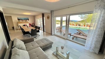 A modern living room with a sofa, a glass coffee table, and a dining area with a table and chairs. There is a sliding glass door leading to a pool area with lounge chairs.