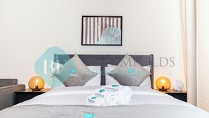 Un lit bien fait, recouvert d’une couette blanche, avec deux oreillers et un ensemble de serviettes pliées. Au-dessus du lit, on aperçoit une œuvre d’art encadrée, une lampe de chevet sur la table de nuit et un luminaire fixé au mur.