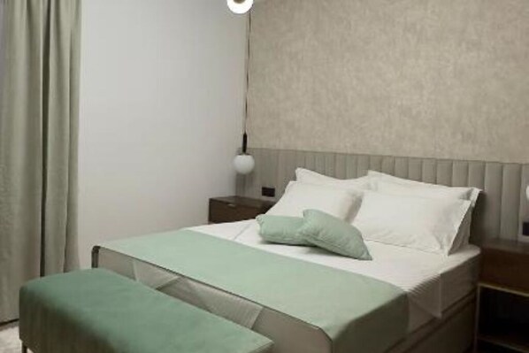A neatly made bed with a green bedspread and pillows, a bedside table with a lamp, and a window with curtains.