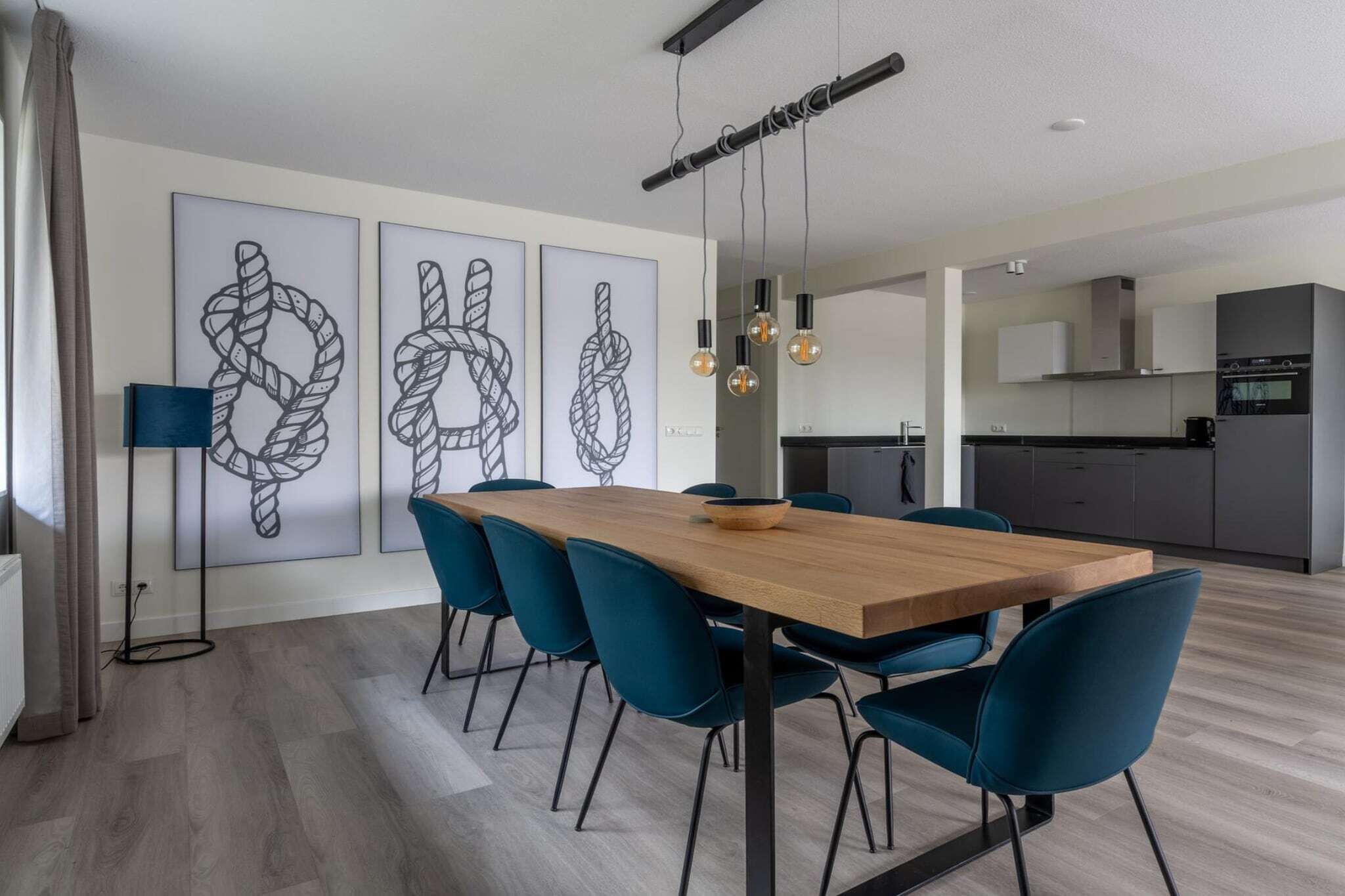 A modern dining area with a wooden table and blue chairs, a kitchen in the background, and abstract art on the wall.