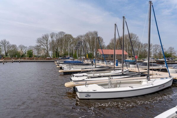 En småbåtshamn med flera segelbåtar förtöjda i stilla vatten, omgiven av träd och en byggnad med ett rött tak.