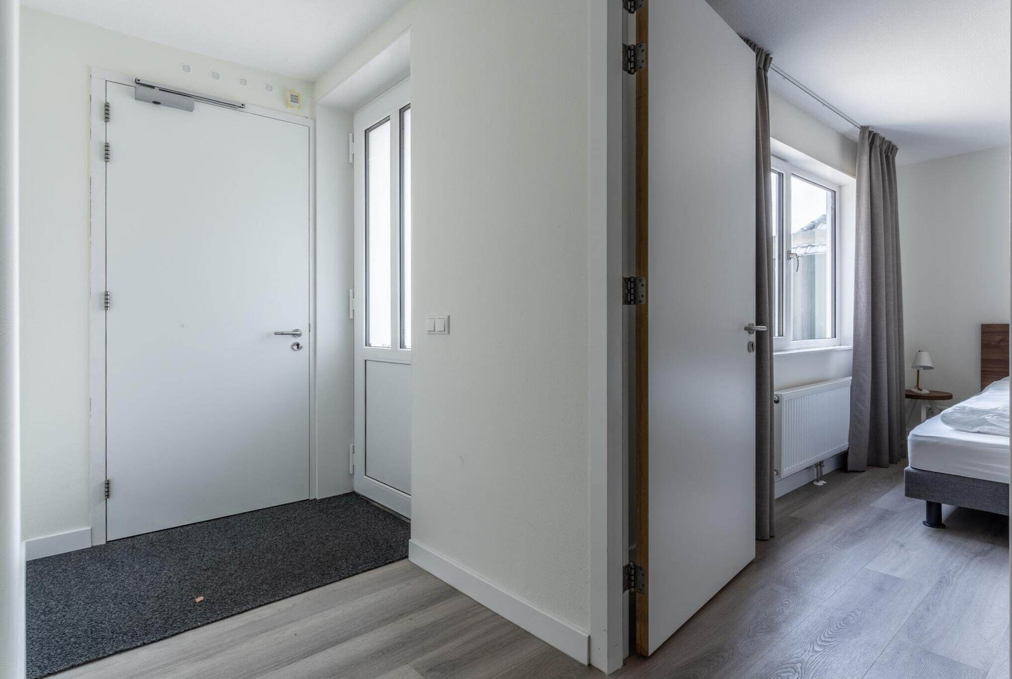 Ein Hotelzimmer mit Bett, Fenster mit Vorhängen, Heizkörper und Tür mit Griff.