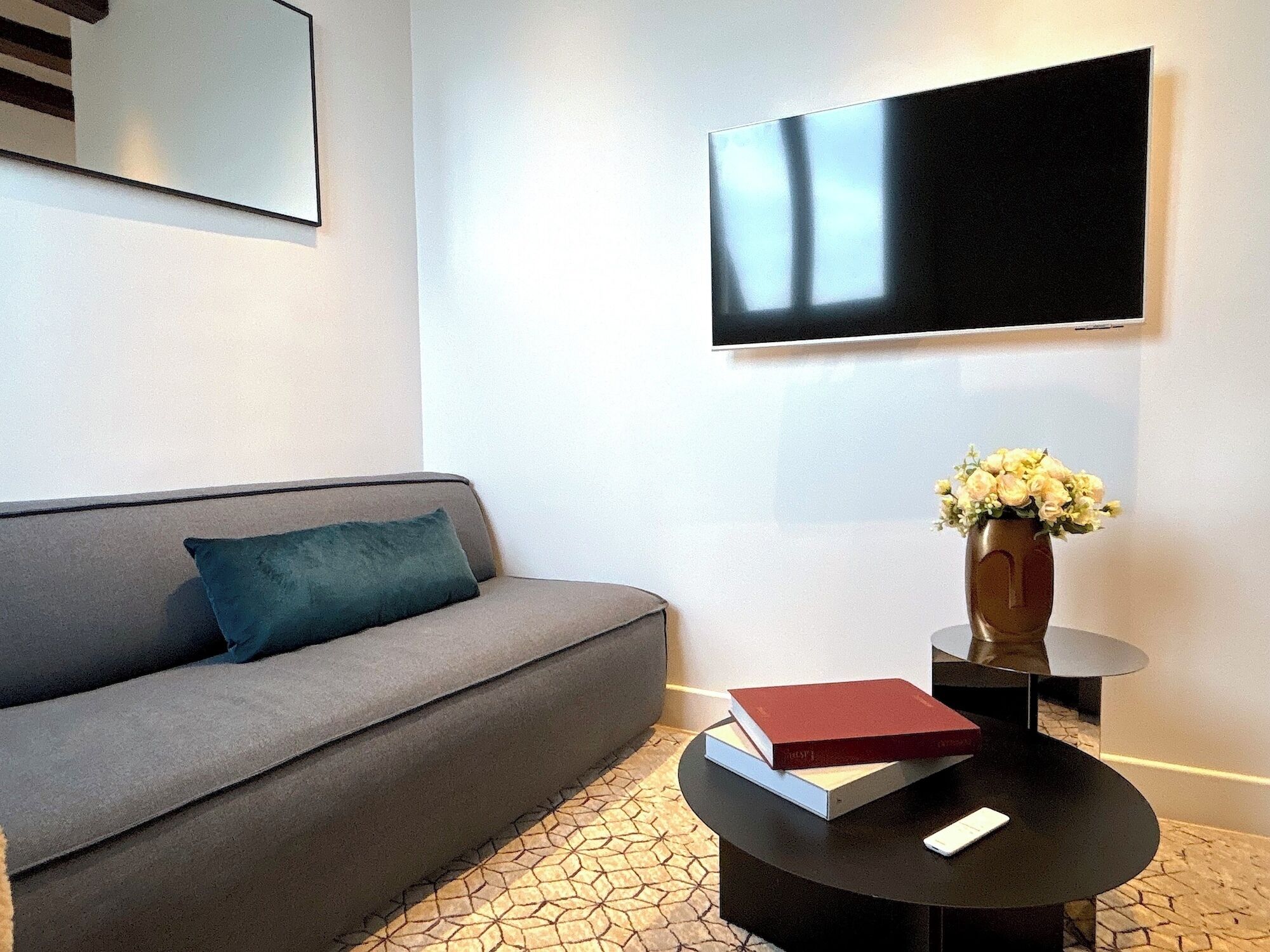 A modern living room with a grey sofa, a round black coffee table, a flat-screen TV mounted on the wall, and a vase with flowers on a side table.