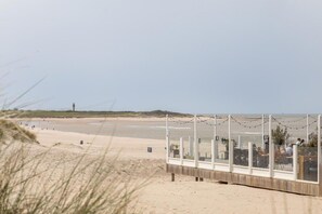 Vista a la playa con muelle de madera, cielo despejado y dunas al fondo.