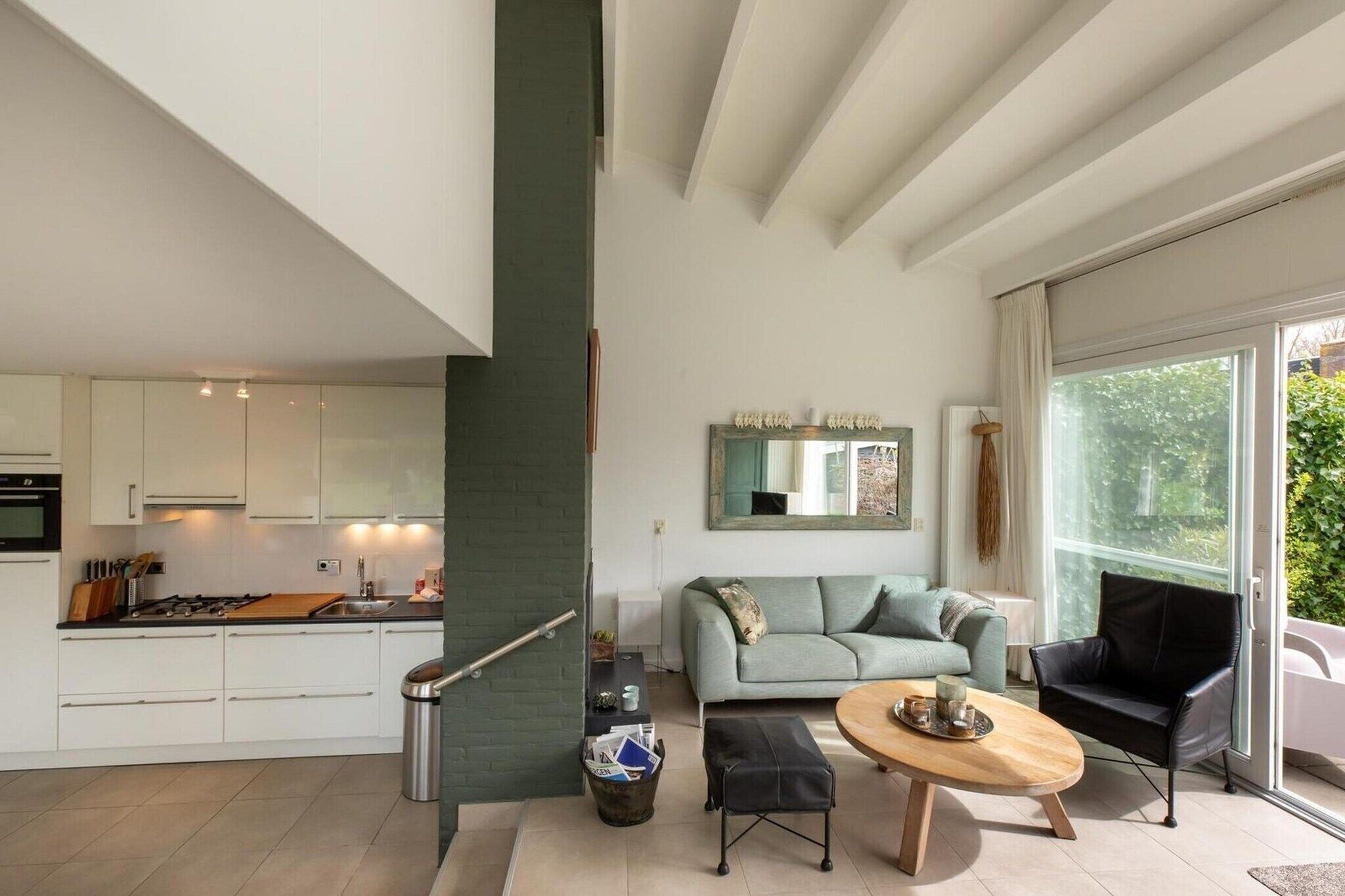 Een moderne keuken met witte kasten, een eethoek met een ronde tafel en een woonkamer met een bank en een zwarte fauteuil.