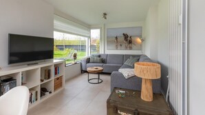 A modern living room with a flat-screen TV, a grey sofa, a coffee table, and a bookshelf. - Holiday Home in Nieuwvliet near the Beach (Nieuwvliet)