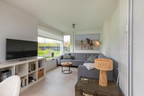 A modern living room with a flat-screen TV, a grey sofa, a coffee table, and a bookshelf. - Holiday Home in Nieuwvliet near the Beach (Nieuwvliet)