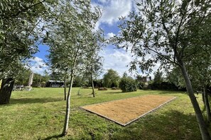 Un espacio al aire libre con césped, una terraza de madera rectangular, árboles y cielo despejado.