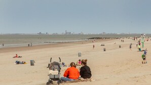 A beach with people, a stroller, and a cityscape in the distance. - Holiday Home in Nieuwvliet near the Beach (Nieuwvliet)