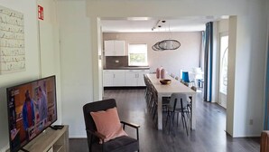 Eine moderne Küche mit weißen Schränken, ein Essbereich mit einem langen Tisch und Stühlen sowie ein Fernseher, der eine Fernsehsendung zeigt.
