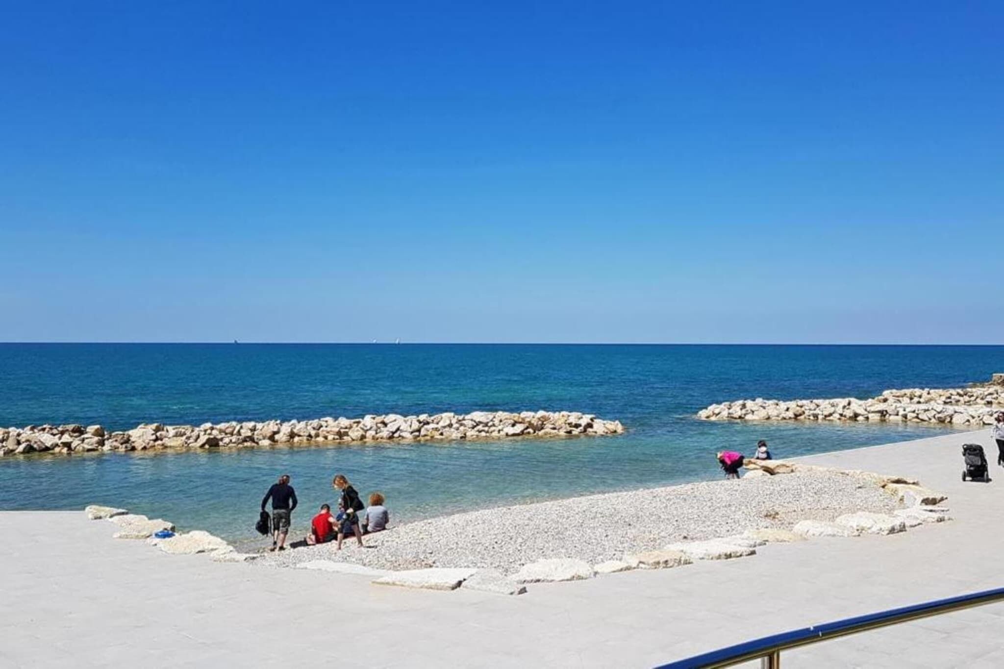 A beach with rocky breakwaters, clear blue water, and people walking along the shore.