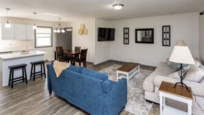 A modern living room with a blue sofa, a grey couch, and a small table. There is a kitchen area with white cabinets and a dining table. - Ankeny Heights - A 4 bedroom and 2 bath home (Ankeny)