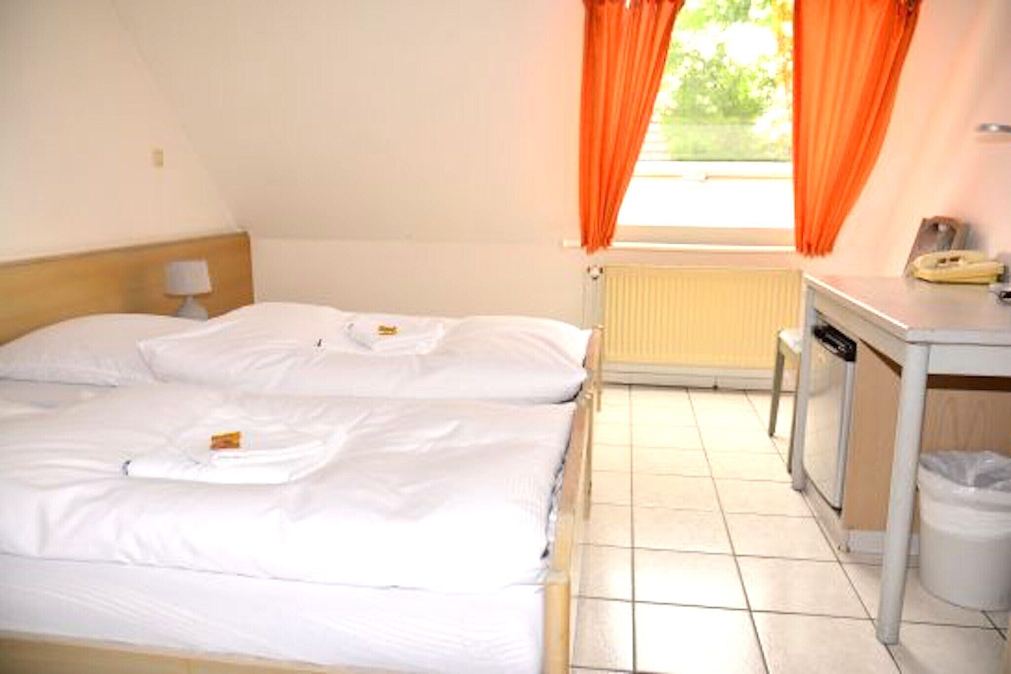Una habitación de hotel con dos camas, una mesita pequeña con un teléfono y una ventana con cortinas anaranjadas.