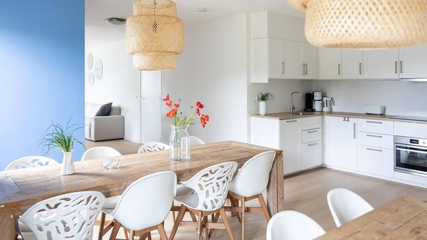 A modern kitchen with white cabinets, a wooden dining table, and white chairs with cut-out patterns. - Holiday Home in Nes near Vleijen Area (Nes)