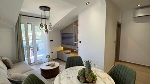 A modern living room with a round marble table, green upholstered chairs, a sofa, and a television in the background.
