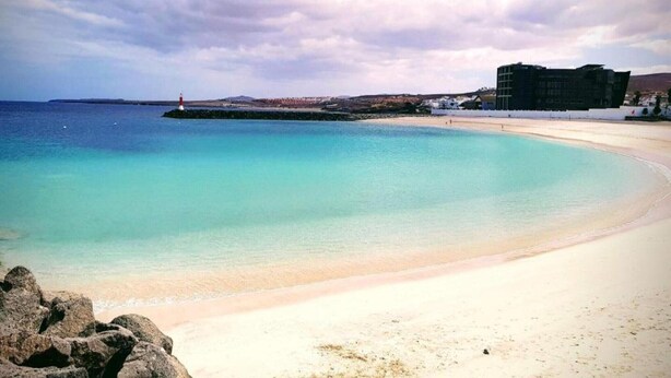 A beach with clear turquoise water, a lighthouse, and a modern building in the background.