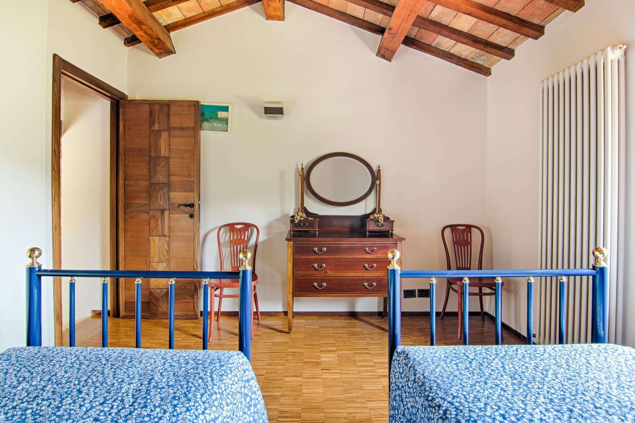 Une pièce avec un plafond en bois, une porte en bois, une commode avec un miroir, deux chaises et un lit recouvert d’une couvre-lit à motifs bleus.