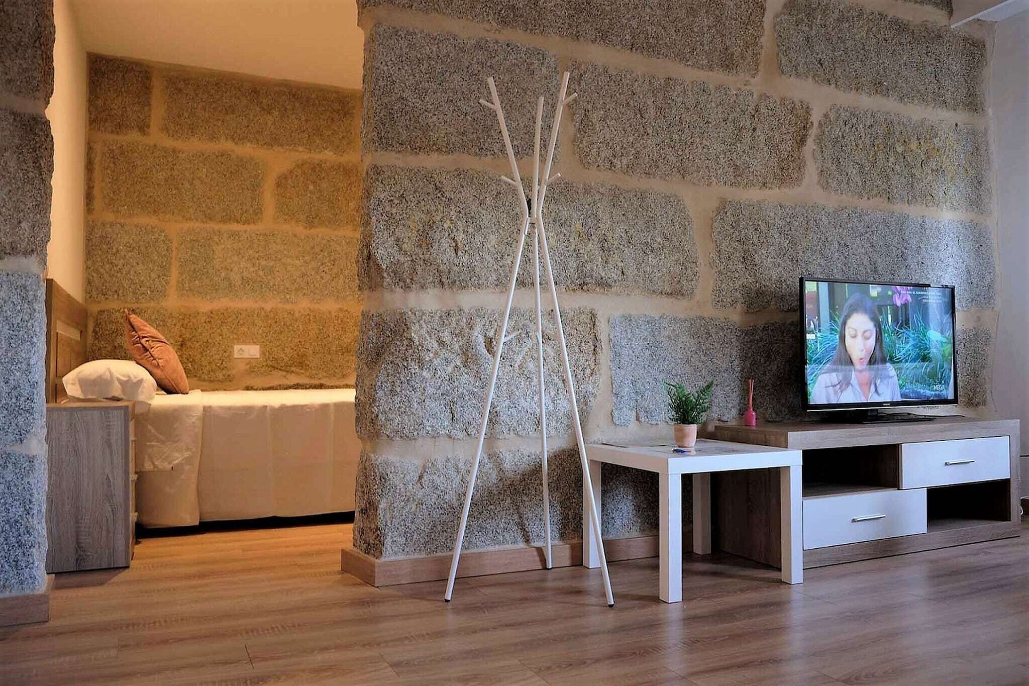 Une pièce avec un mur en pierre, un lit avec un matelas blanc et un oreiller marron, un bureau blanc avec une télévision et une chaise blanche.