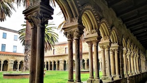 Un patio con una serie de columnas y arcos, un área verde y un edificio con ventanas al fondo.