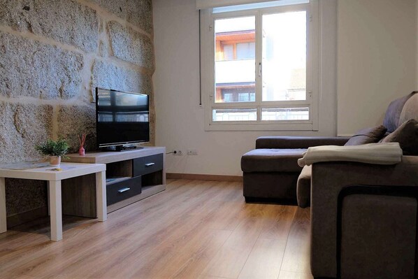 Una sala de estar con pared de piedra, un televisor de pantalla plana sobre un mueble blanco, un sofá gris y una ventana con vistas a edificios.