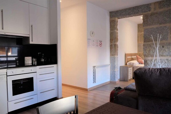 Una cocina moderna con armarios blancos y horno empotrado, pared de piedra, cama con cabecero de madera y una mesita de noche pequeña.