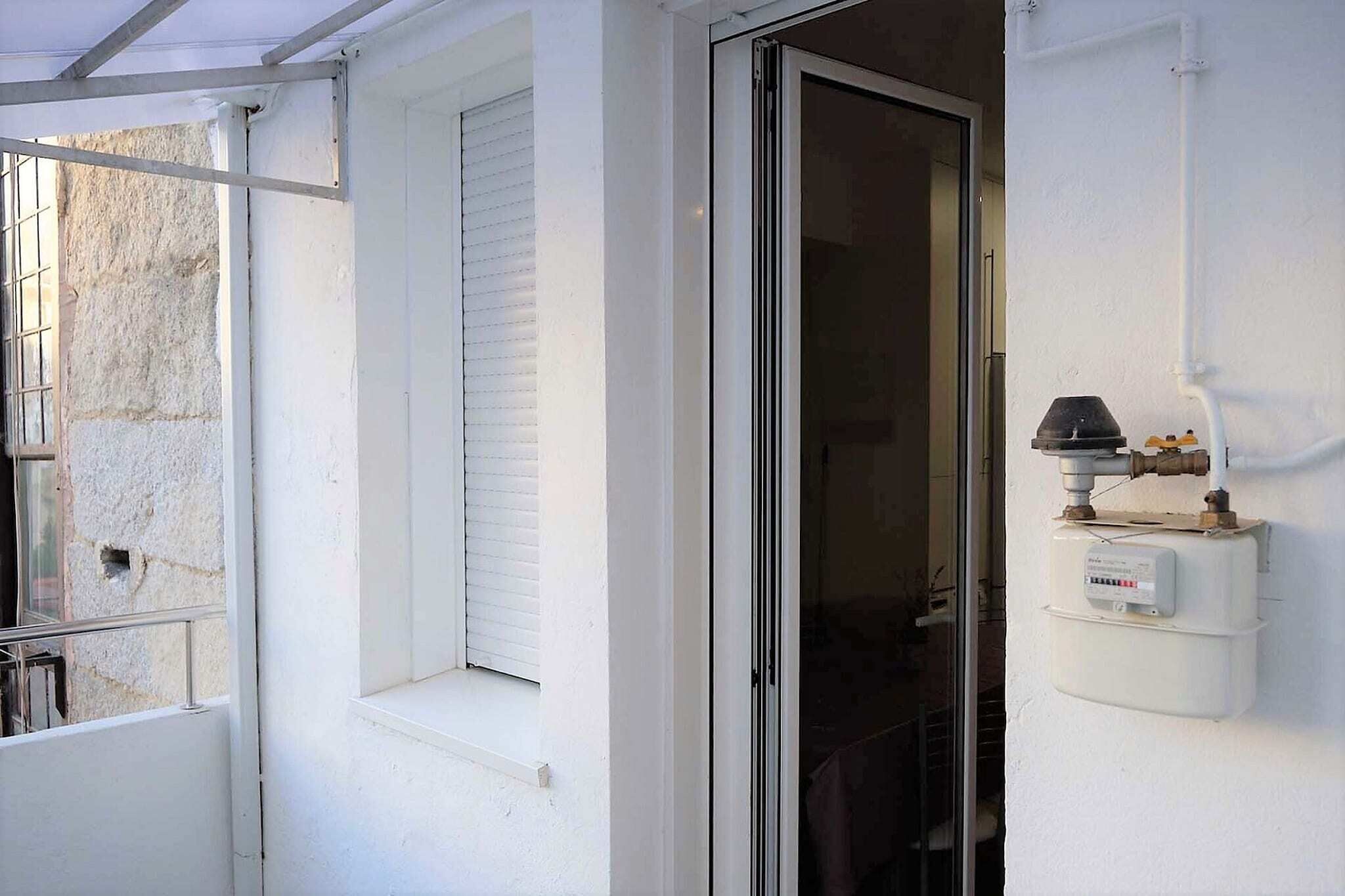 Pared exterior blanca con una ventana y un divisor de vidrio corredero. Se observa un medidor de agua adherido a la pared.