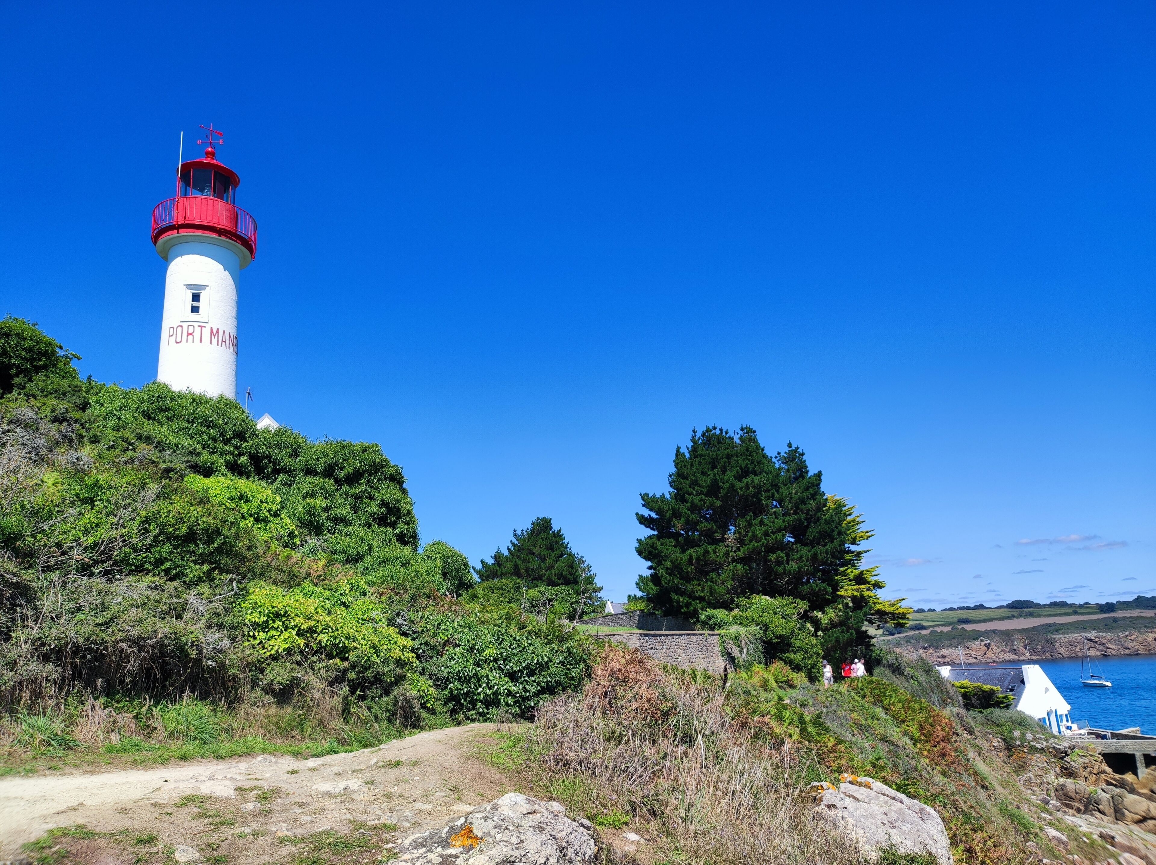 A lighthouse named "Port Man" with a red top, surrounded by greenery and a clear blue sky.
