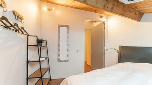 A bedroom with a bed, a wooden floor, a black metal shelf with a plant, a mirror, and a door leading to another room.