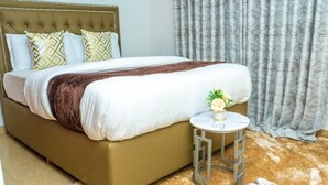 A bed with a brown headboard, white bedding, and patterned pillows. A small round table with a vase of flowers is beside the bed. - Lux suites Jimka Apartments Kisumu (Kisumu)