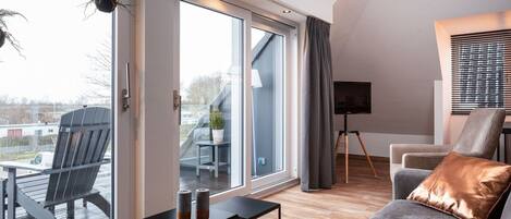 Un salon moderne avec un canapé gris, une petite table d’appoint noire et une grande fenêtre donnant sur un balcon et les bâtiments environnants.