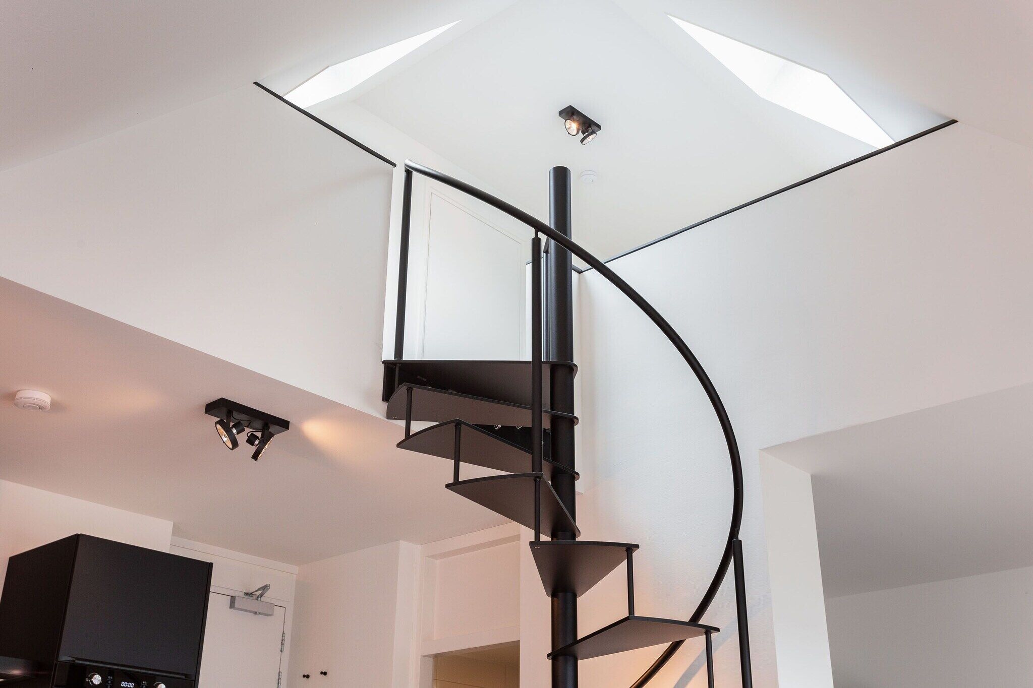 Una escalera espiral moderna con diseño minimalista, un techo plano con iluminación empotrada y un mueble de entretenimiento negro al fondo.