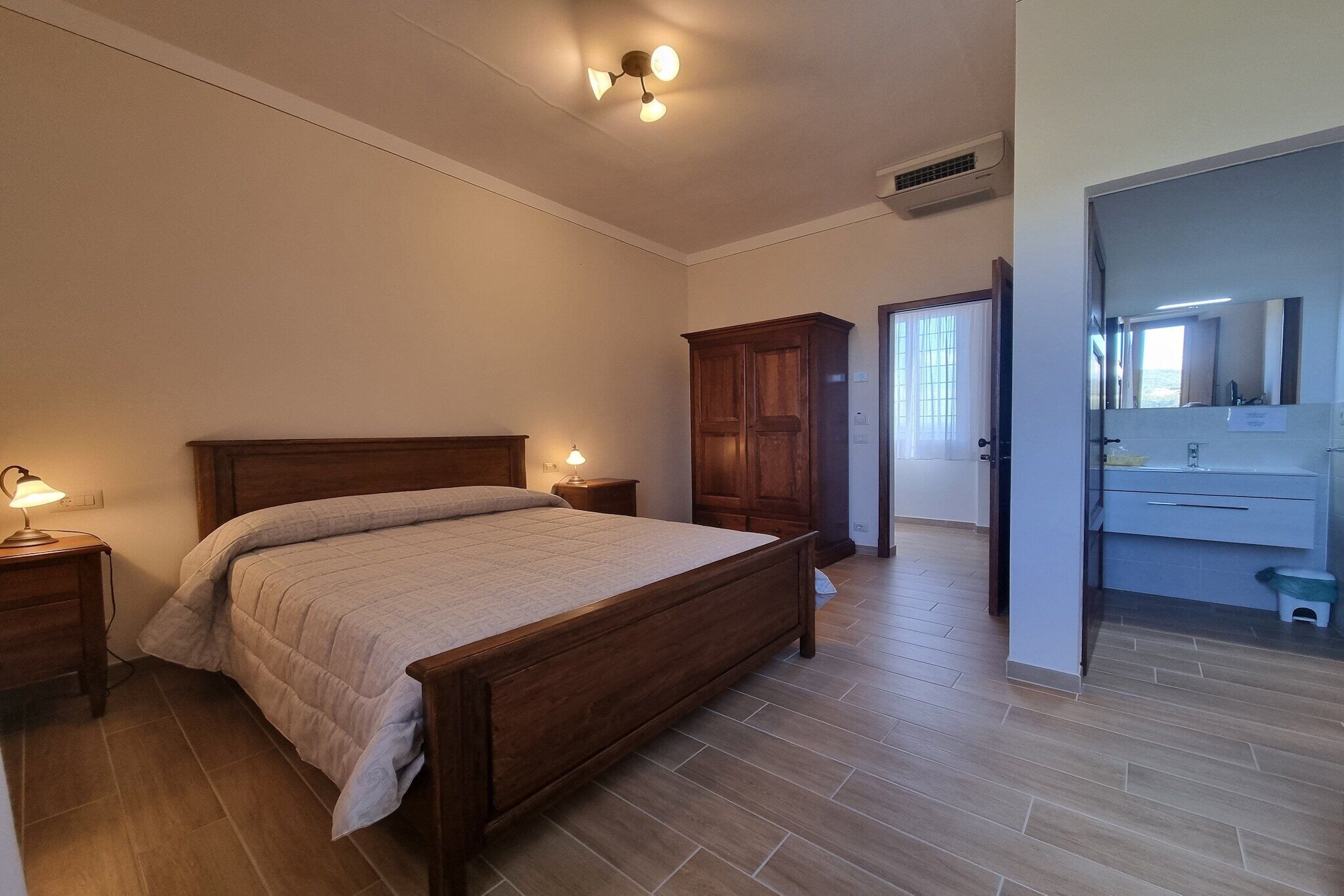 Un dormitorio con cama de madera, lámparas de noche, un armario y un baño visible a través de una puerta abierta.