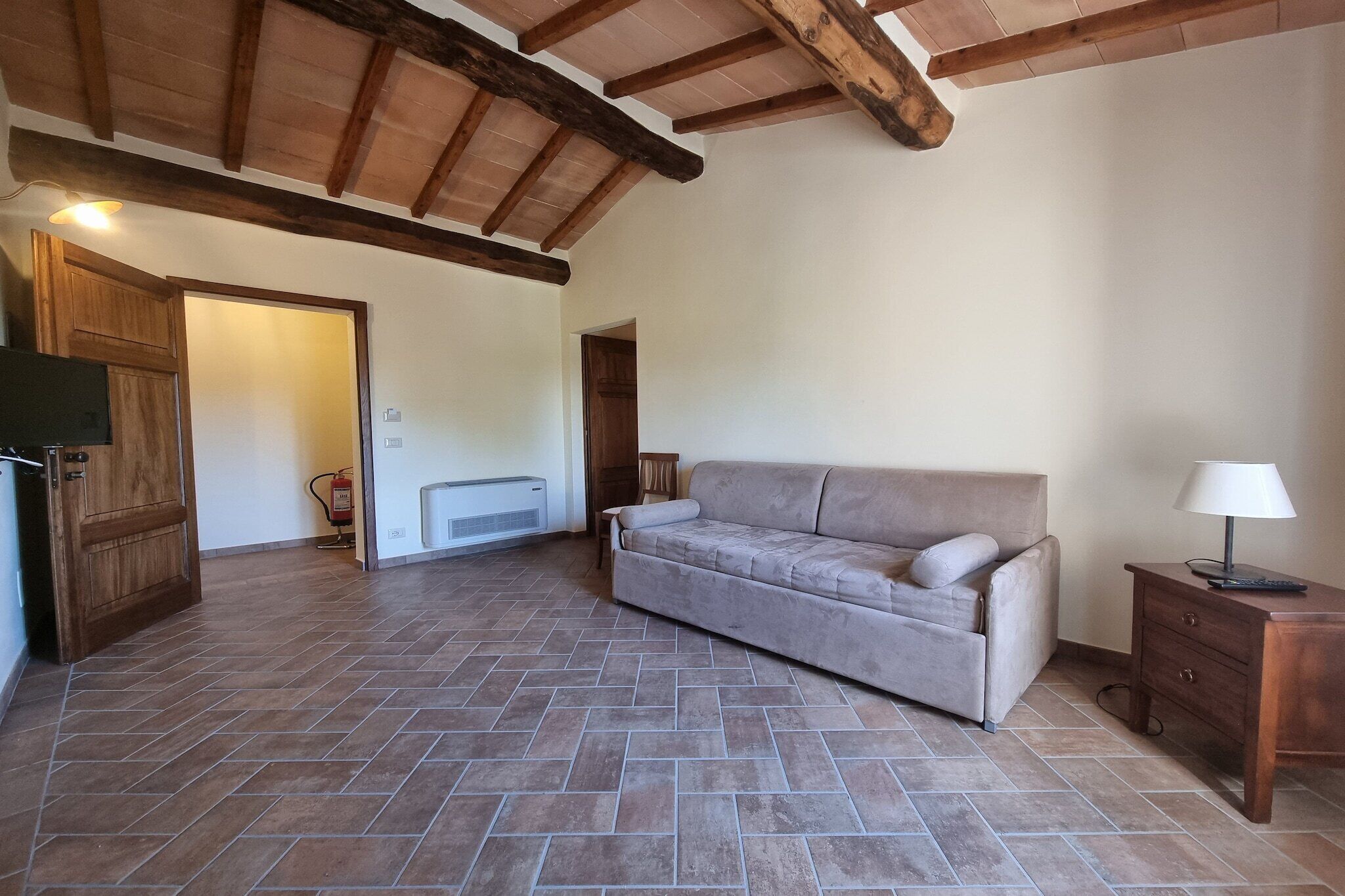 A spacious living area with a beige sofa, a wooden cabinet, and a small table with a lamp.