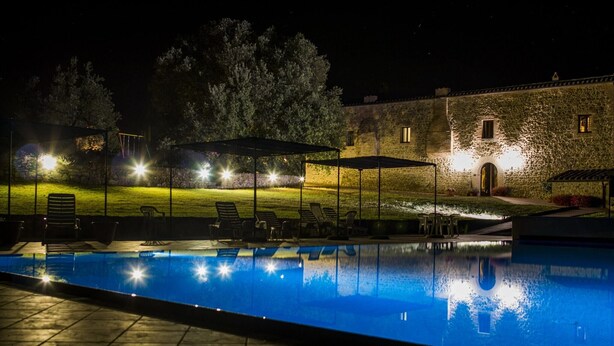 A nighttime swimming pool area with lounge chairs and umbrellas, surrounded by stone buildings and trees.