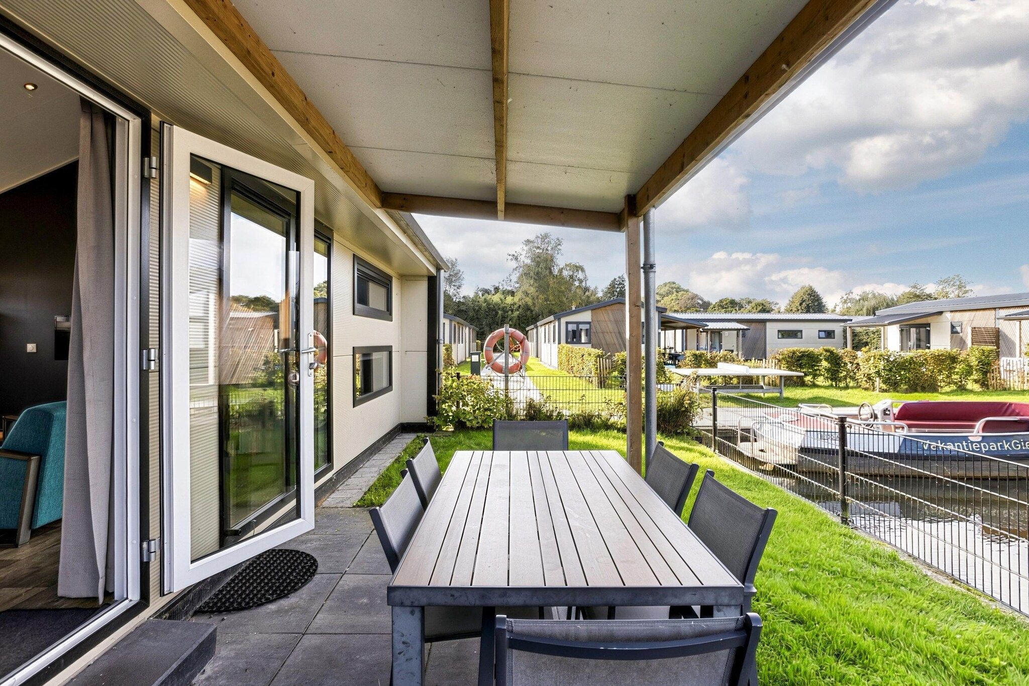 Eine überdachte Terrasse mit Tisch und Stühlen, ein Glastür-Eingang und Blick auf eine Wohngegend mit Häusern und einem Boot.
