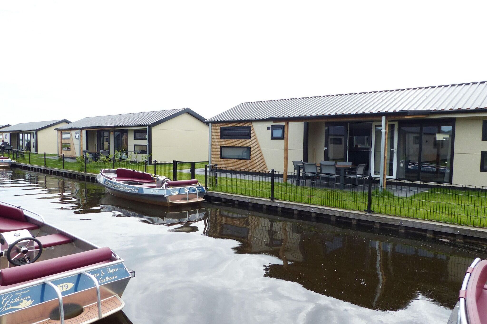 A row of modern, single-story buildings with large windows and a flat roof, adjacent to a calm canal with boats docked.