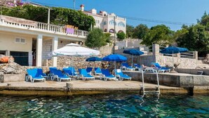 A poolside area with blue lounge chairs and umbrellas, a stone building with a balcony, and clear blue water.