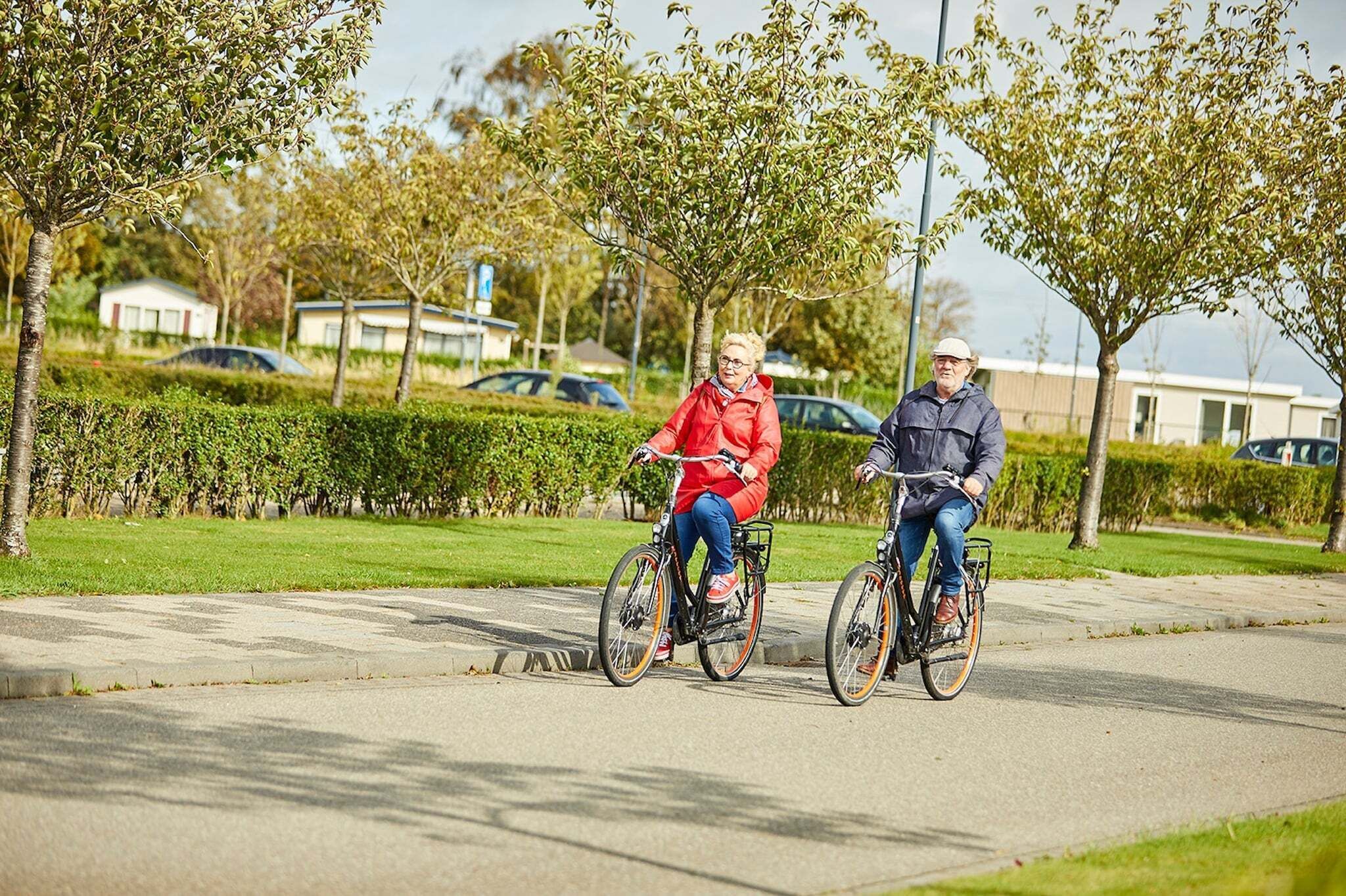 Dos personas paseando en bicicleta por un camino pavimentado en un parque, con árboles y un edificio al fondo.