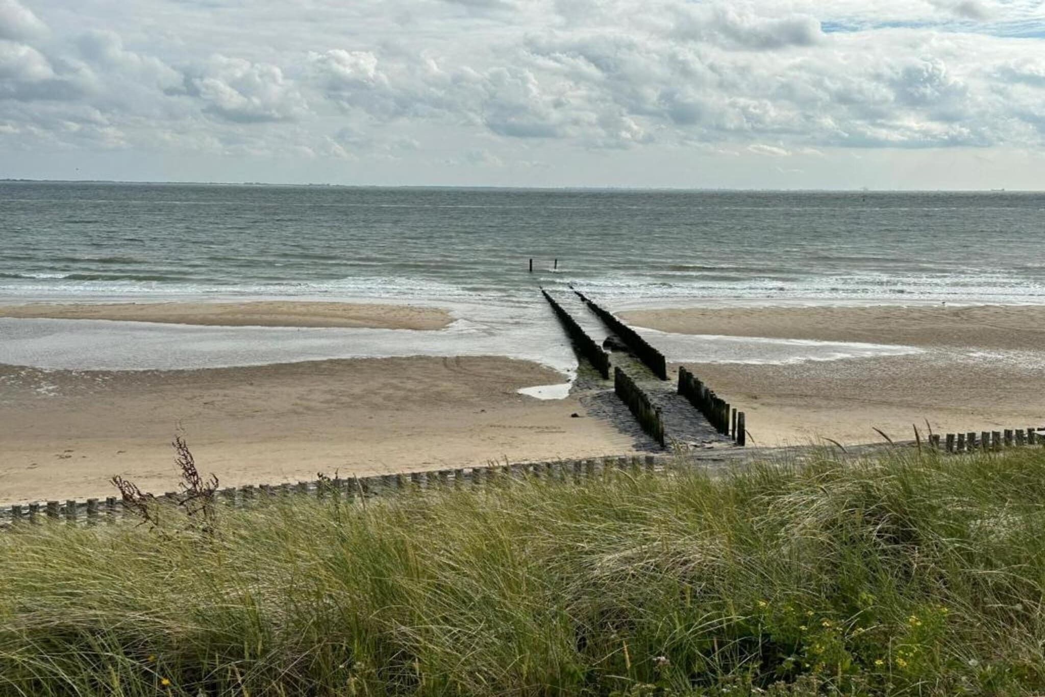 Ein Strand mit einer hölzernen Steg, der ins Meer führt, und Dünen im Vordergrund, sowie ein bewölkter Himmel.