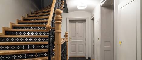 Una escalera con escalones de madera y barandillas negras, que conduce a una puerta blanca al final de un pasillo con suelo estampado.