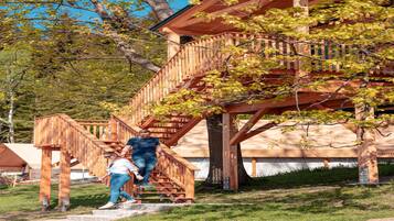 Un escalier en bois mène à une terrasse en bois d’où l’on peut admirer un arbre et un bâtiment en arrière-plan.