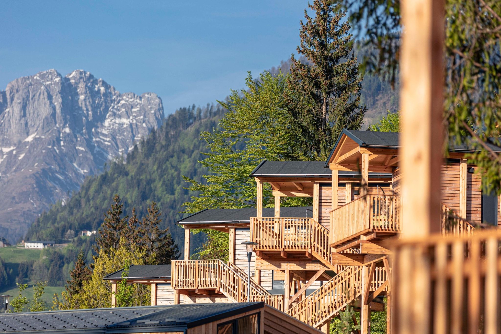 A wooden cabin with multiple levels and balconies, set against a mountainous backdrop.