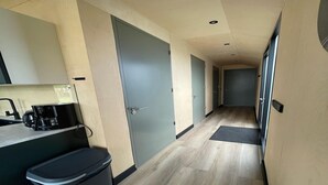Un couloir étroit avec un plancher en bois clair, une machine à café, une poubelle et une porte au fond.