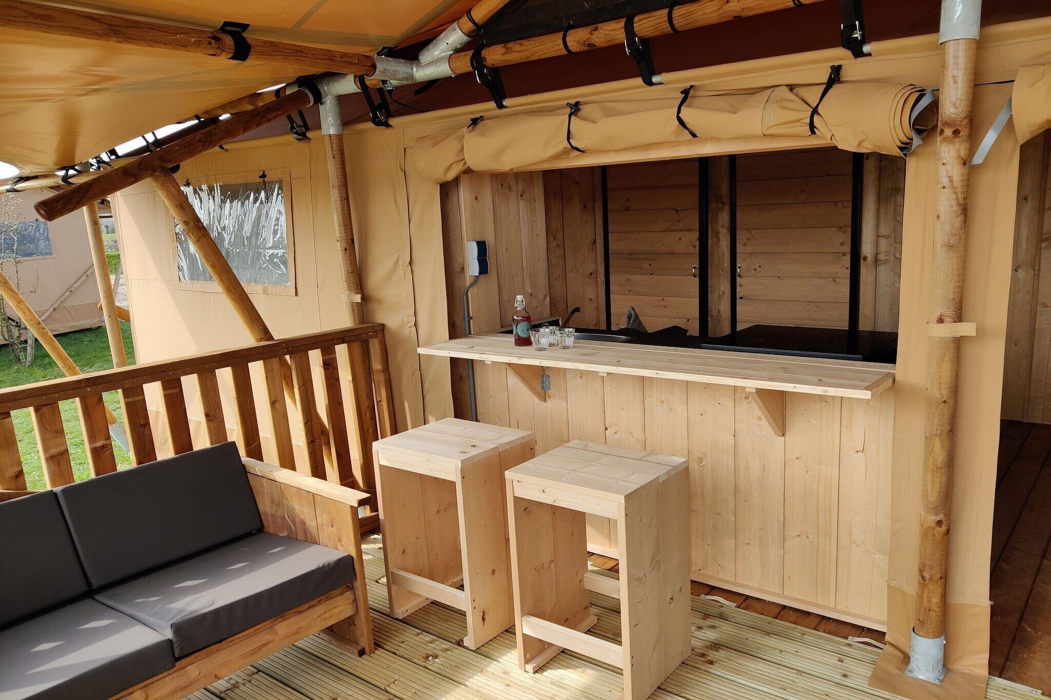 Une terrasse en bois avec un banc et une petite table, un espace abrité avec une fenêtre, et une vue sur l’extérieur.